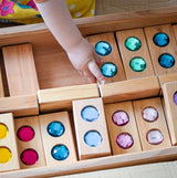 Child's hand placing a Bauspiel Color Track gem block back in the wooden tray · view 3