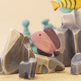 Wooden discus fish painted dusty pink, beside a pale rock · view 1
