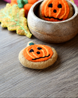 Round orange felt cookie with a frazzled pumpkin face needle-felted into the surface · view 1